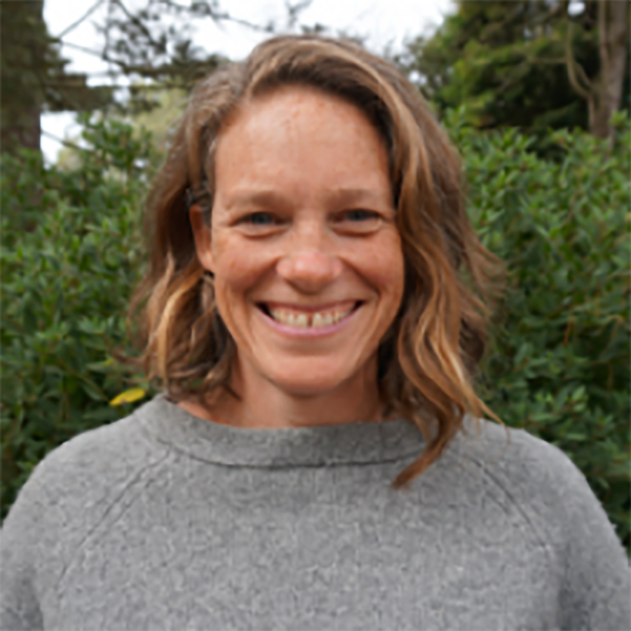 A woman with blonde, shoulder length hair and a gray sweater smiles at the camera. There is a line of trees in the background.