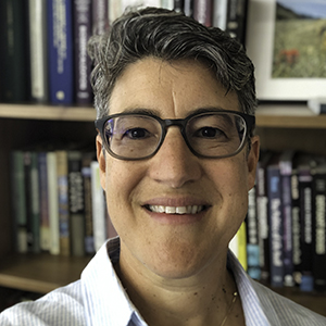 A short haired woman with glasses is smiling in front of a bookcase.