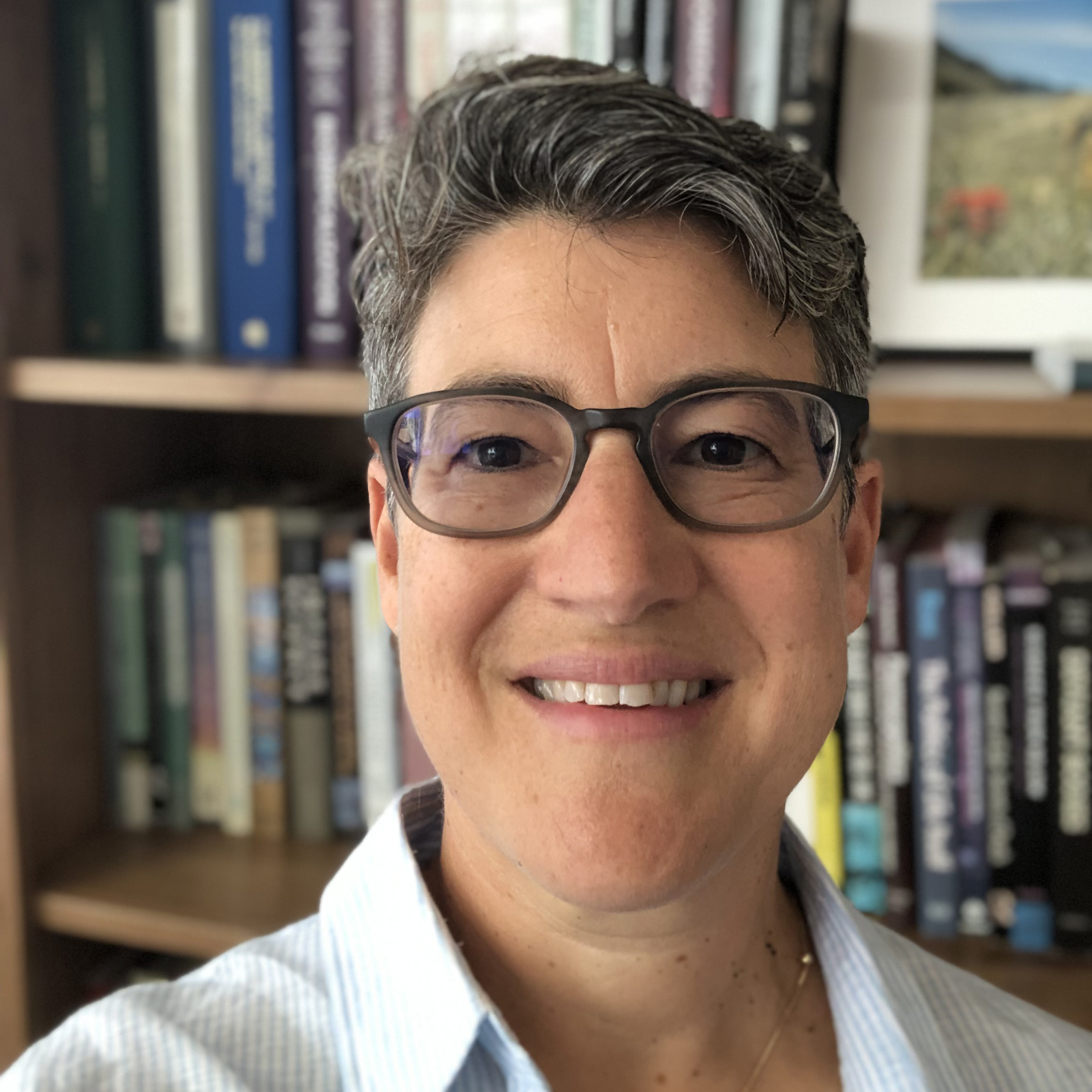 A short haired woman with glasses is smiling in front of a bookcase.