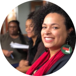 A Black woman wearing a red scarf and black jacket with green, black, white, and red earrings looks toward her left toward the camera, smiling.