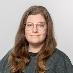 A woman wearing wire frame glasses and long, dark blonde hair from the shoulders up. Her shirt is dark green.