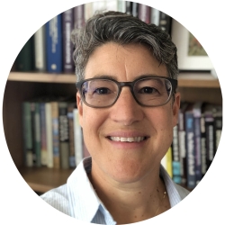 A woman with short, salt and pepper hair and glasses smiles into the camera. She is in front of a wall of books housed in a wooden bookcase.