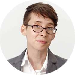 A woman with short hair and glasses from the shoulders up is wearing a white button up under a gray blazer.