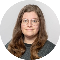 A woman with long dark-blonde hair and glasses from the shoulders up. She wears a forest-green crewneck shirt and is in front of a solid gray background.