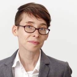 A headshot of a white woman with short hair and glasses. They are wearing a white button-up and a gray blazer.