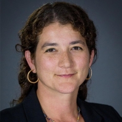 A light-skinned woman from the shoulders up. She has long, dark, curly hair tied back. She wears gold hoop earrings and a black blazer. She is smiling at the camera.