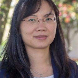 An Asian woman with long dark hair and wire framed glasses. She wears a navy blue blazer and a striped boatneck underneath. She stand in front of dappled sunlight and leaves of a tree.